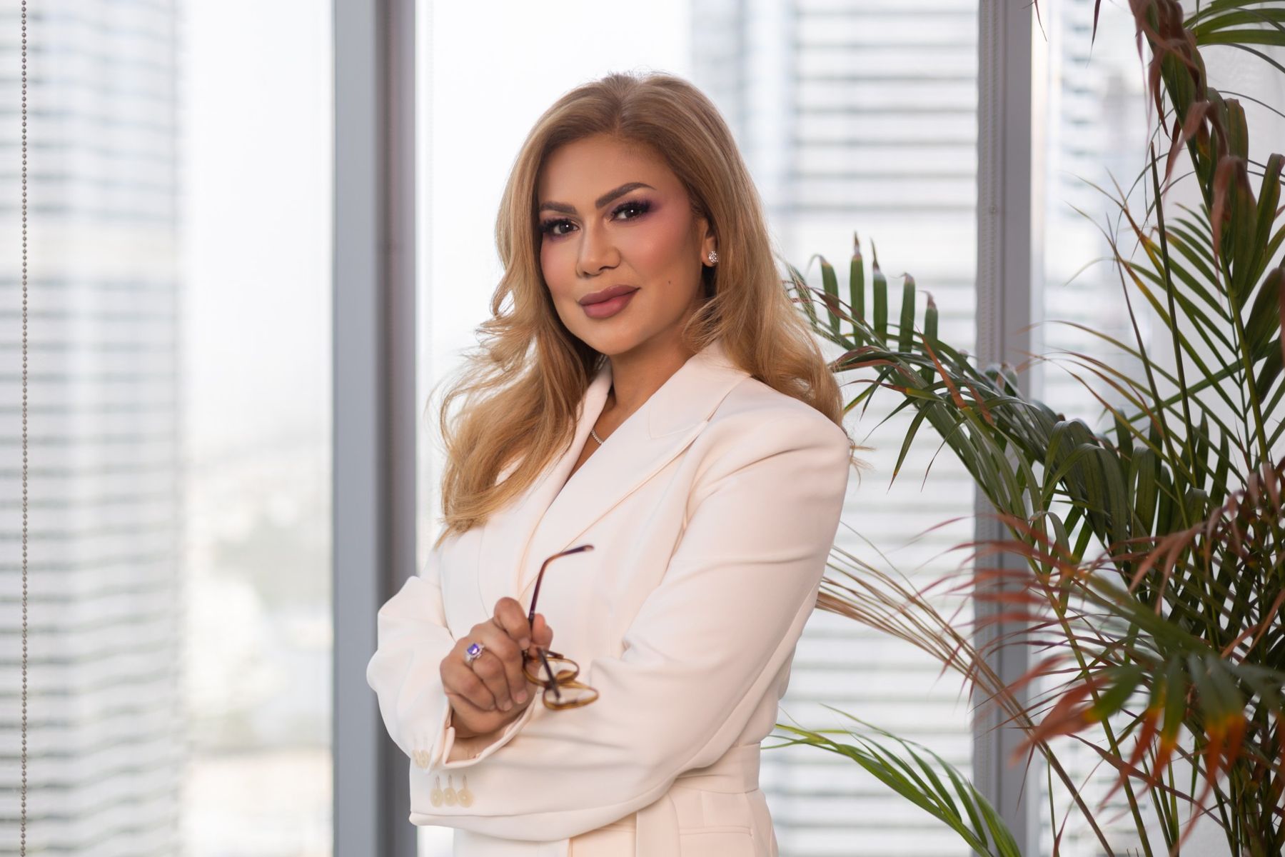 Wardah Harharah in a cream suit, holding her glasses, by an office window